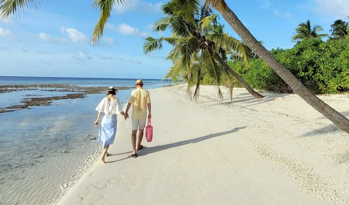 A couple strolls hand in hand along a serene tropical beach under swaying palm trees, with clear blue skies and gentle ocean waves