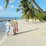 A couple strolls hand in hand along a serene tropical beach under swaying palm trees, with clear blue skies and gentle ocean waves