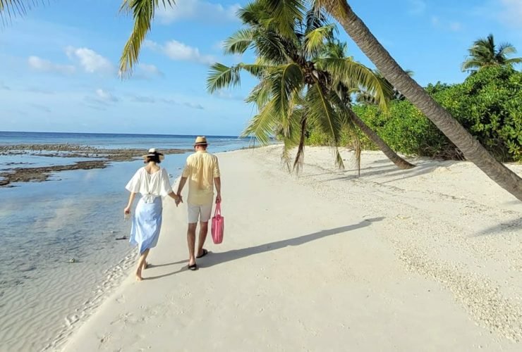 A couple strolls hand in hand along a serene tropical beach under swaying palm trees, with clear blue skies and gentle ocean waves